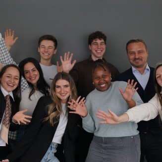 A diverse group of eight people poses joyfully in front of a gray backdrop, raising their hands and smiling. They are dressed in a mix of business and casual attire, with some wearing blazers while others wear sweaters or t-shirts. The lively energy and friendly expressions suggest a fun and celebratory moment, possibly for a team or group photo.
