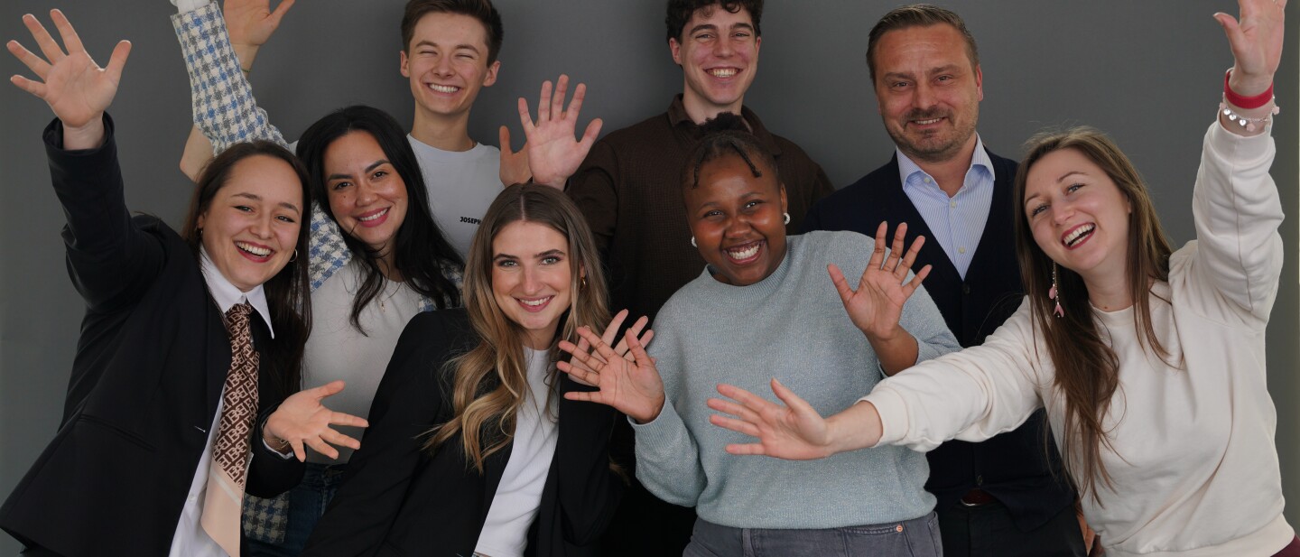 A diverse group of eight people poses joyfully in front of a gray backdrop, raising their hands and smiling. They are dressed in a mix of business and casual attire, with some wearing blazers while others wear sweaters or t-shirts. The lively energy and friendly expressions suggest a fun and celebratory moment, possibly for a team or group photo.