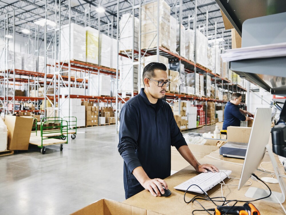 A male warehouse worker checking orders at computer workstation in warehouse