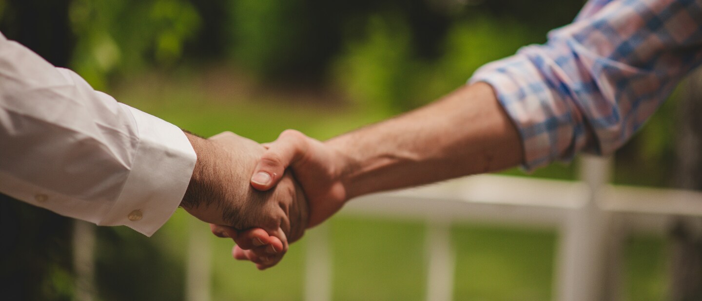 Two people shake hands outdoors, one wearing a white dress shirt and the other in a pastel plaid shirt. The handshake is centered in the frame, symbolizing agreement or partnership, with a blurred green garden background.