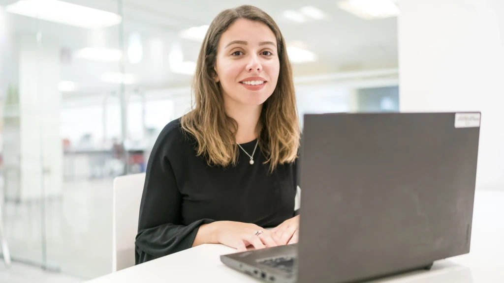 Eine junge Frau sitzt vor ihrem Laptop und lächelt in die Kamera.