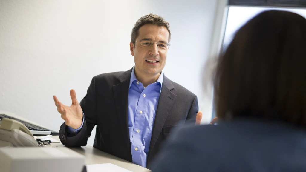 Ein Mann sitzt am Tisch und spricht mit einer Kollegin.