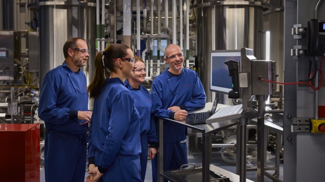 A group of four professionals wearing matching blue protective suits and safety glasses stand together in an industrial facility, smiling and engaged in a discussion. They are gathered around a workstation with a laptop and a control panel, which appears to be monitoring or operating machinery. The background features large stainless steel tanks and complex piping, suggesting a high-tech manufacturing or laboratory environment.