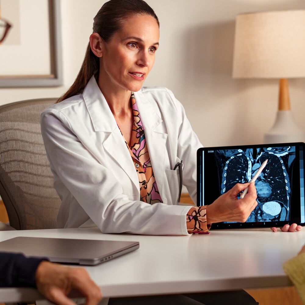 A female doctor in a lab coat pointing to an xray of lungs on a tablet while explaining something to two seated patients.